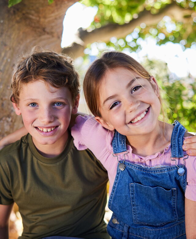 Due bambini sono seduti all’aperto sotto un albero, sorridono alla telecamera e si tengono amichevolmente abbracciati.
