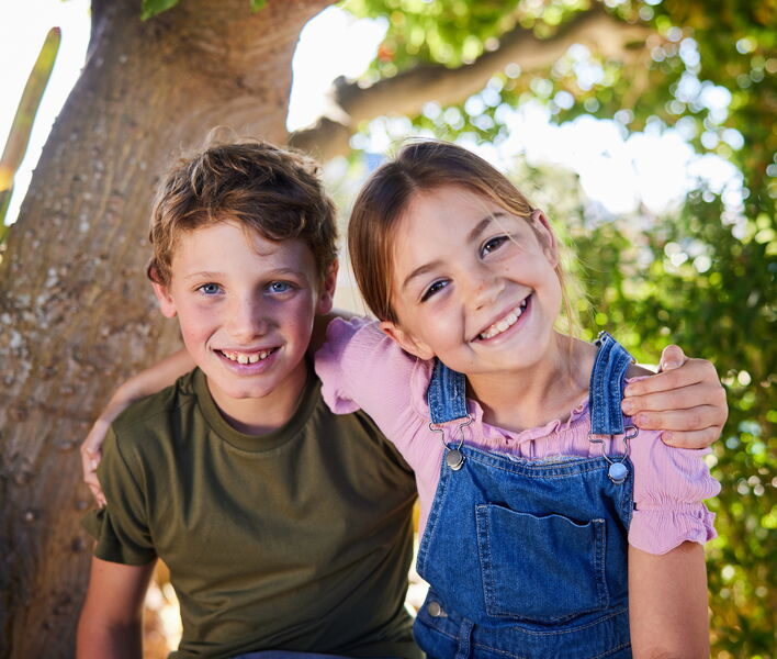 Zwei Kinder sitzen draussen unter einem Baum, lächeln in die Kamera und haben die Arme freundschaftlich umeinander gelegt.