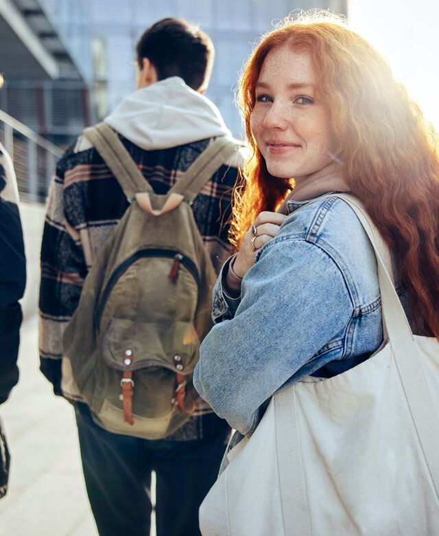 Eine junge Frau mit Tasche blickt im Gehen lächelnd in die Kamera, während sie gemeinsam mit anderen jungen Menschen einen Weg entlanggeht.