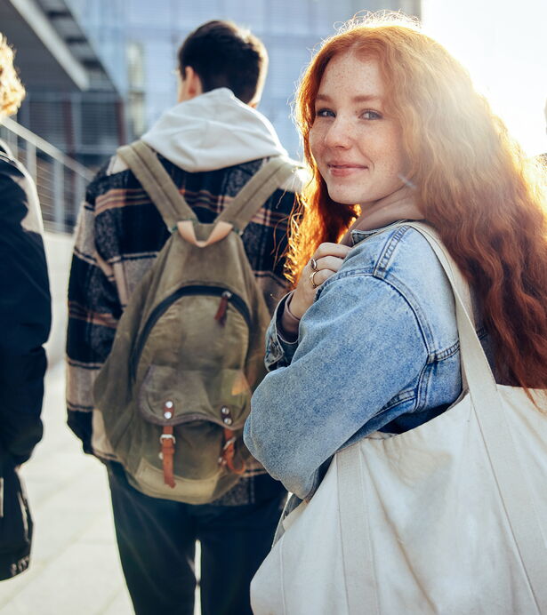 Una giovane donna con una borsa sorride alla telecamera mentre cammina lungo un percorso insieme ad altri giovani.