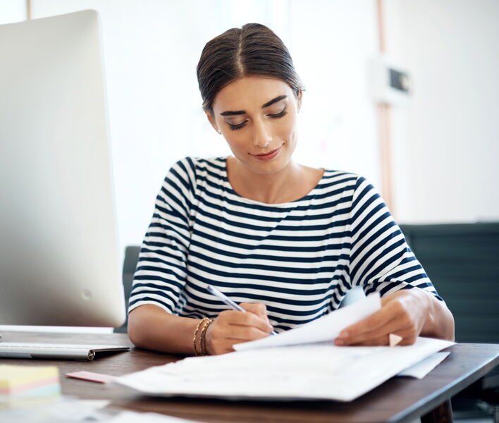 Eine Person sitzt am Tisch, arbeitet am Laptop und hält dabei ein Dokument in der Hand.