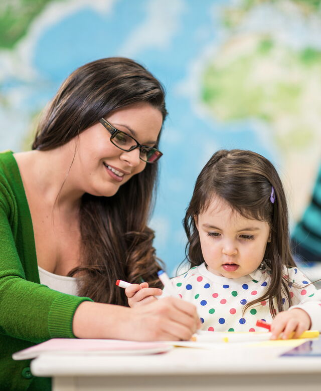 Eine Betreuungsperson unterstützt ein kleines Mädchen beim Zeichnen an einem Tisch.