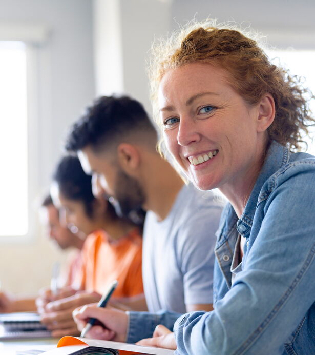 Eine Frau sitzt in einem Seminarraum, lächelt in die Kamera und schreibt.