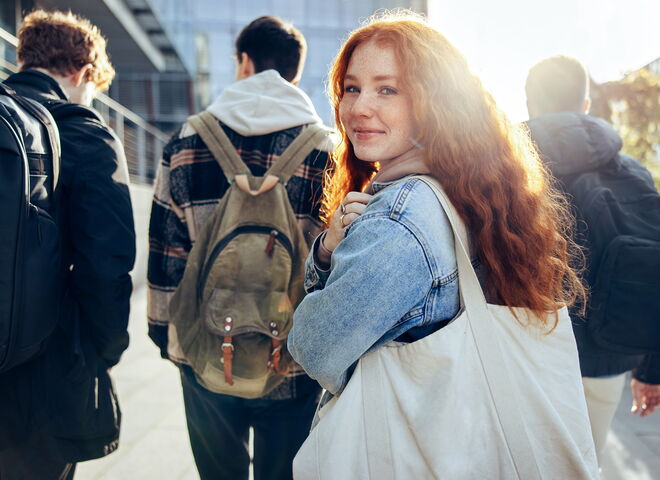 Eine junge Frau mit Tasche blickt im Gehen lächelnd in die Kamera, während sie gemeinsam mit anderen jungen Menschen einen Weg entlanggeht.