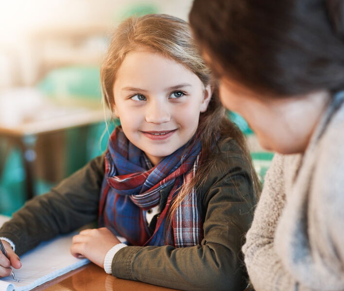 Ein Mädchen sitzt an einem Tisch, schreibt in ein Heft und blickt lächelnd zu einer Betreuungsperson neben ihr.
