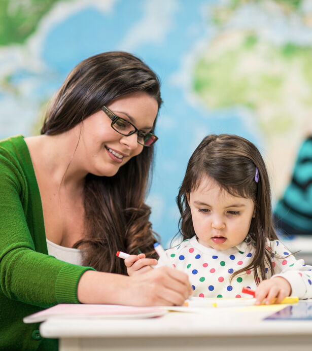 Una persona di accompagnamento aiuta una bambina a disegnare a un tavolo.